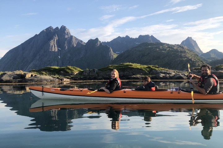 Three people kayaking on a calm lake with rocky mountains in the background under a clear sky.