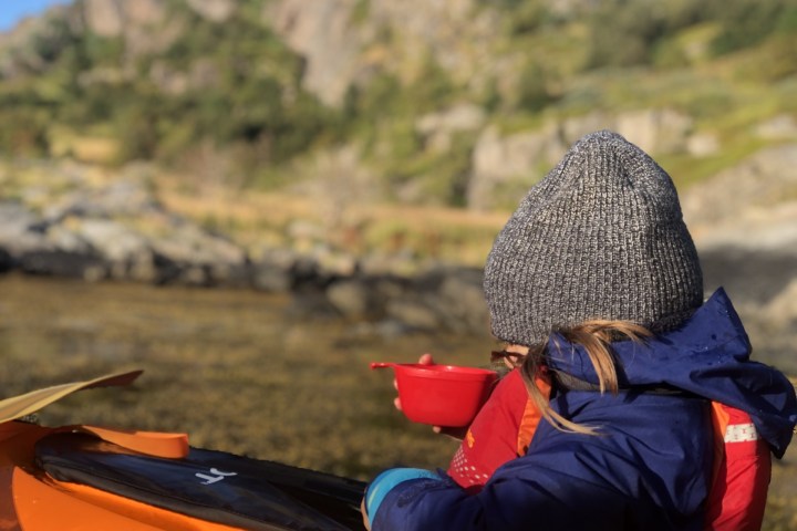 Child in winter clothing holding a red bowl near a kayak with blurred mountains in the background.