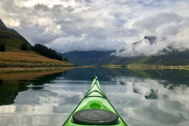 View from a kayak on a calm lake, surrounded by mountains and cloudy sky.