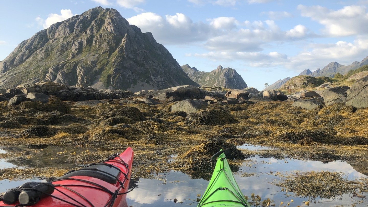 Red and green kayaks on calm water with mountains and cloudy sky in the background.