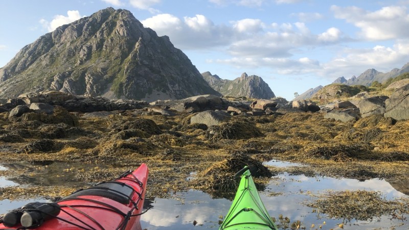 Red and green kayaks on calm water with mountains and cloudy sky in the background.