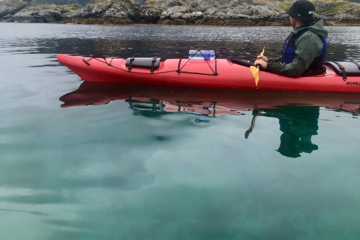 Person kayaking on clear water near rocky shore and misty mountains.