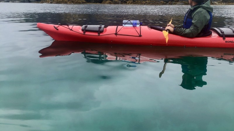 Person kayaking on clear water near rocky shore and misty mountains.