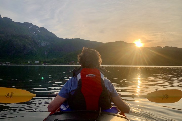 Person kayaking on calm water at sunset with mountains in the background.