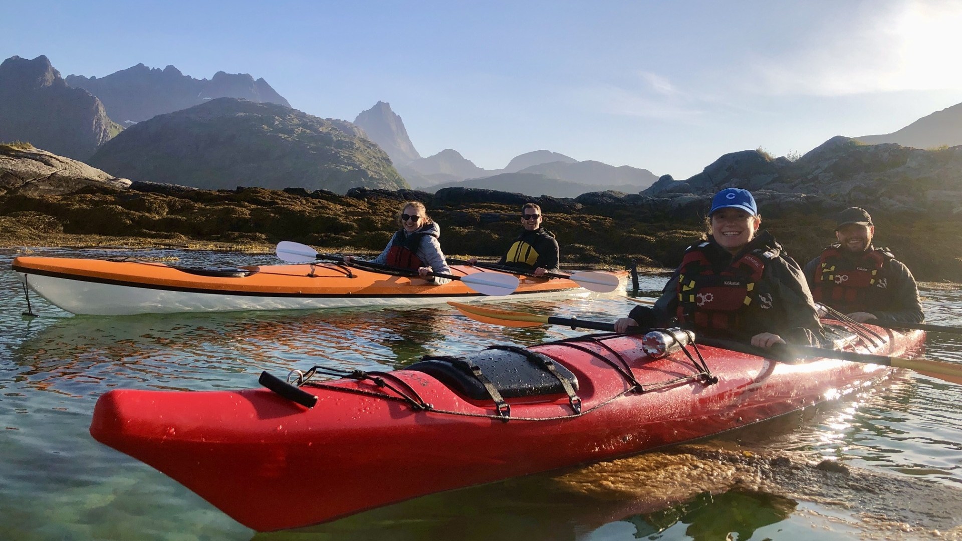 Three people kayaking in clear water with mountains in the background.