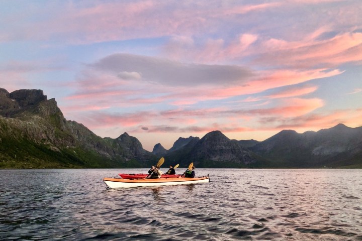 Two kayakers on a lake with mountain backdrop under a pink and blue sunset sky.
