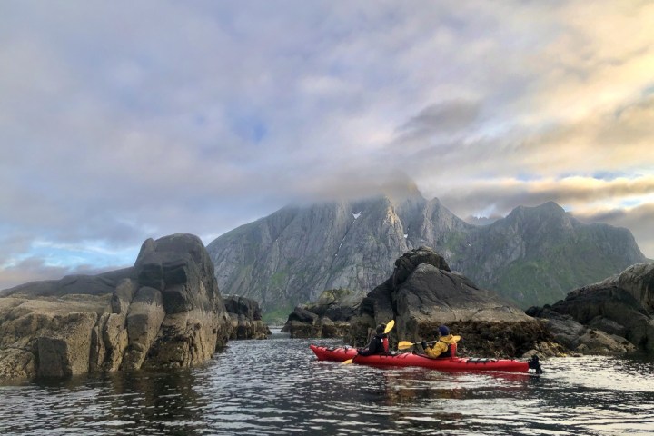 Two people kayaking in red kayaks between rocky formations with mountains under a partly cloudy sky.