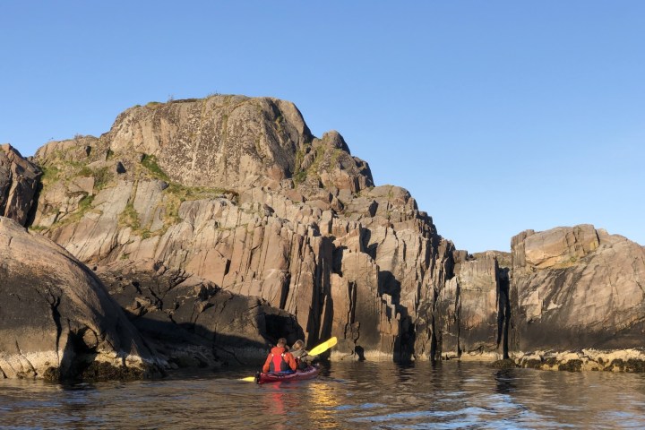 Two people kayaking near rocky cliffs under a clear blue sky.