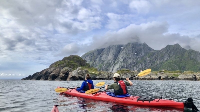 Two kayakers paddle near rocky islands, with mountains and cloudy sky in the background.