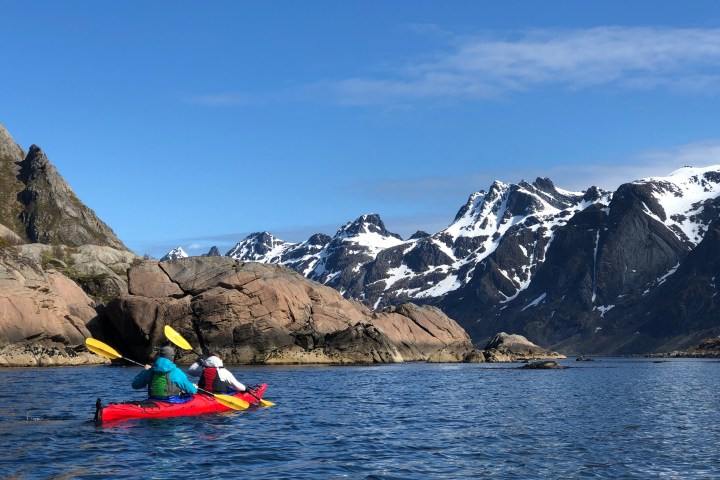 Two kayakers in red kayak on blue lake with snow-capped mountains and rocky shore under clear sky.
