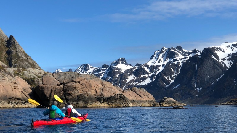 Two kayakers in red kayak on blue lake with snow-capped mountains and rocky shore under clear sky.
