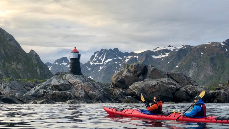 Two kayakers paddle near a rocky shore with a small lighthouse and snow-capped mountains in the background.