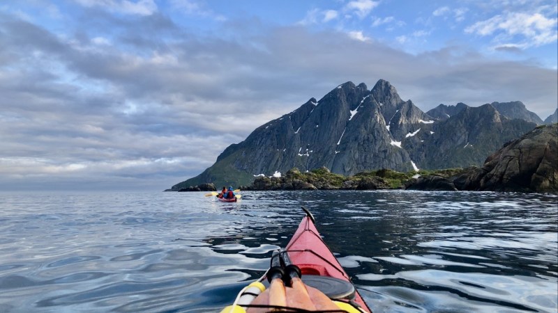 Kayakers on calm water, mountain backdrop, cloudy sky.