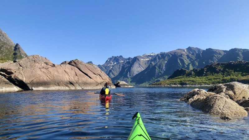 Person in yellow jacket kayaking between rocky islands with mountains in the background under a clear blue sky.