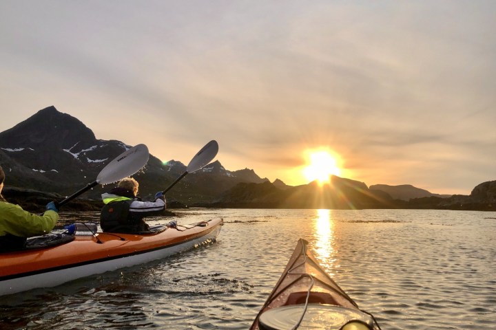 Two kayakers paddle towards the sun setting over distant mountains on a calm lake.