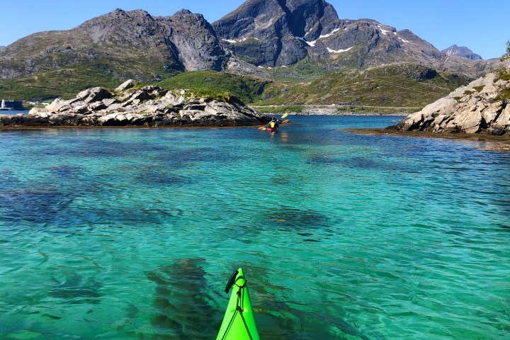 Green kayak in clear blue water with rocky mountains under blue sky.
