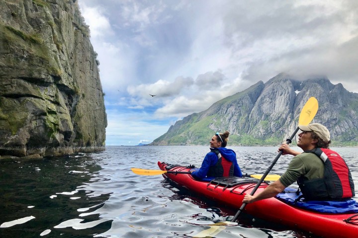 Two people kayaking near a rocky cliff with mountains in background under a cloudy sky.