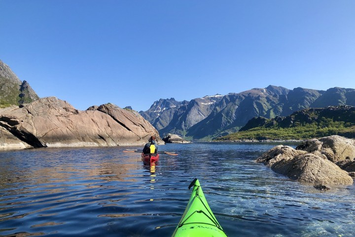 Person in a kayak paddling towards rocky mountains under a clear blue sky.