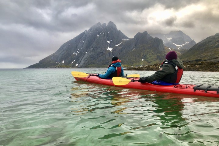 Two people kayaking on a calm sea with cloudy mountains in the background.