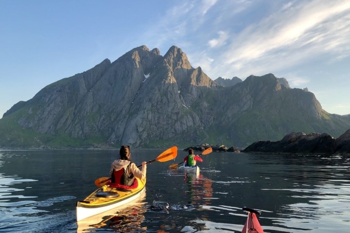 Three kayakers paddling toward rocky mountains under a clear blue sky.