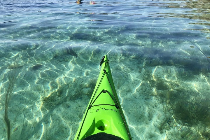 Clear water view from a green kayak with two people in a red kayak ahead, surrounded by mountains.