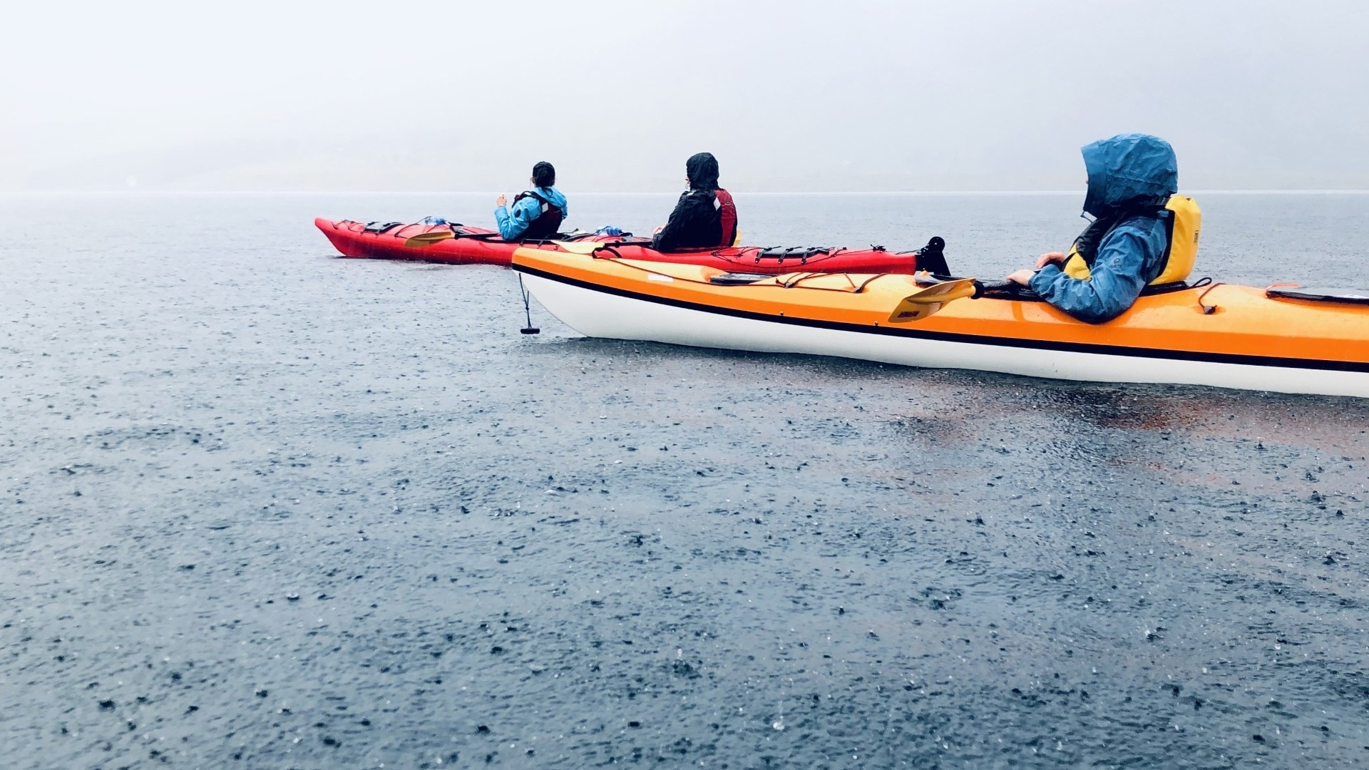 Three people kayaking on a rainy, misty lake with overcast skies.