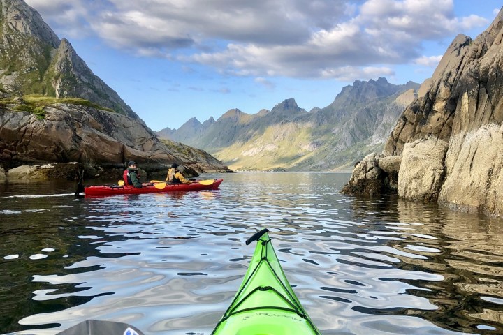 View from a green kayak on a calm fjord with mountains and a red kayak in the distance.