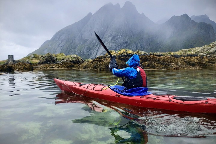 Person kayaking in red kayak near rocky shore and mountains under cloudy sky.