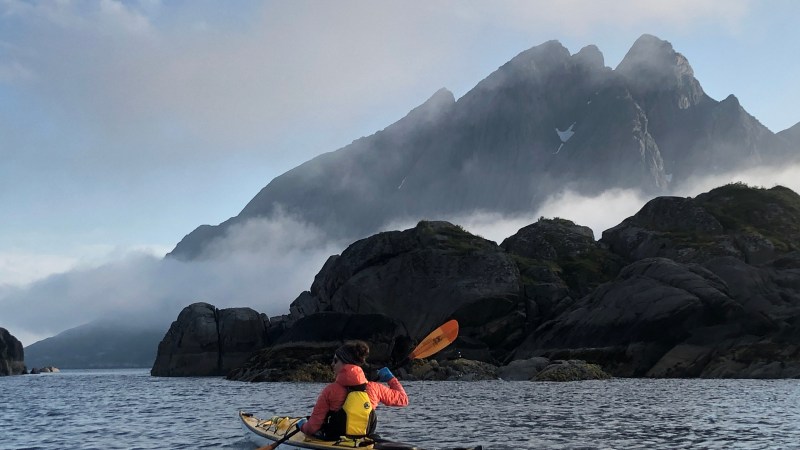 Person kayaking on calm water near rocky shore with misty mountains in background.