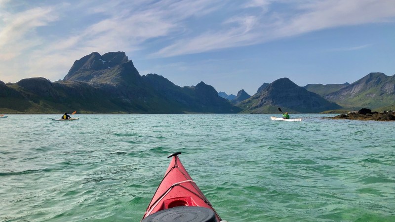 View from a red kayak on a calm sea with mountains and other kayakers in the background.