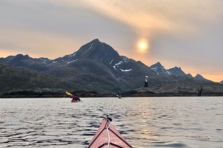 Kayakers on water with mountains, lighthouse, and sun in background under a cloudy sky.