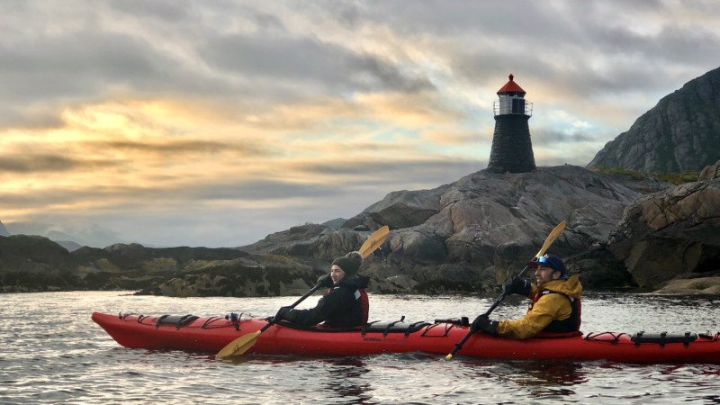Two people kayaking near a lighthouse on rocky shore at sunset.