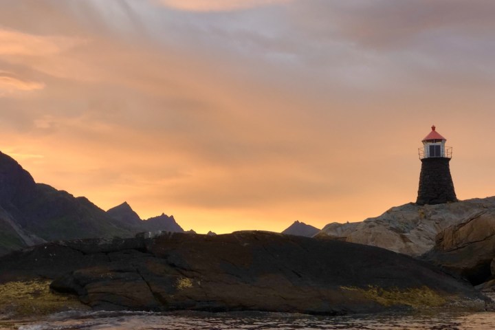 Lighthouse on rocky coast at sunset with colorful sky and mountain silhouette.
