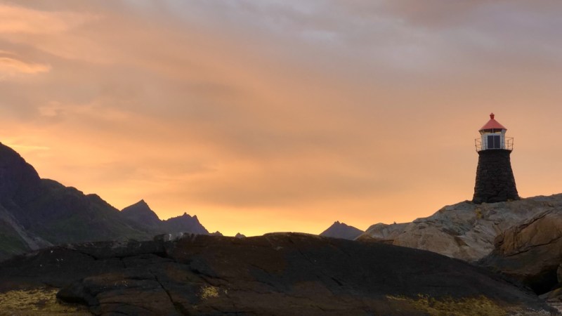 Lighthouse on rocky coast at sunset with colorful sky and mountain silhouette.