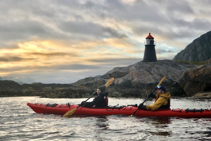 Two people kayaking near a rocky shore with a lighthouse at sunrise.