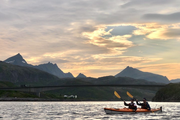 Two people kayaking on a calm lake with mountains and bridge in the background during sunset.