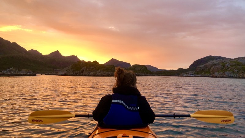 Person kayaking on calm water at sunset with mountains in the background.