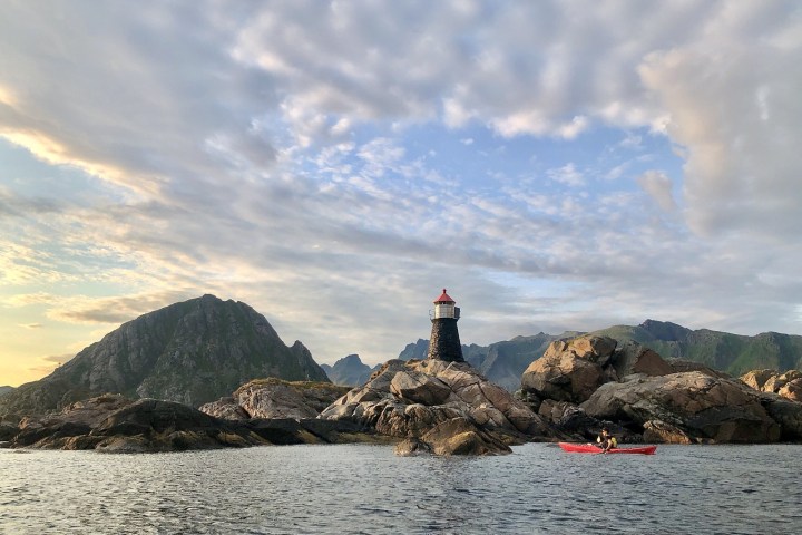 Kayaker near rocky island with small lighthouse and mountains under a cloudy sky.