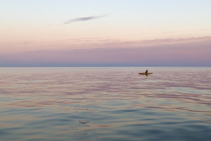 Person in kayak on calm sea during sunset with pastel sky and clouds.