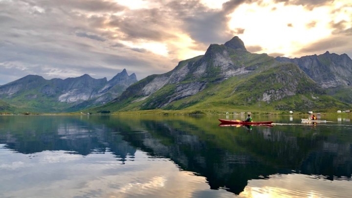 Kayakers on a calm lake with mountains and a dramatic sky in the background, reflecting in the water.