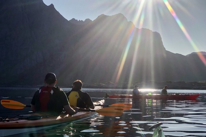 People kayaking on calm water with sun rays and mountains in the background.