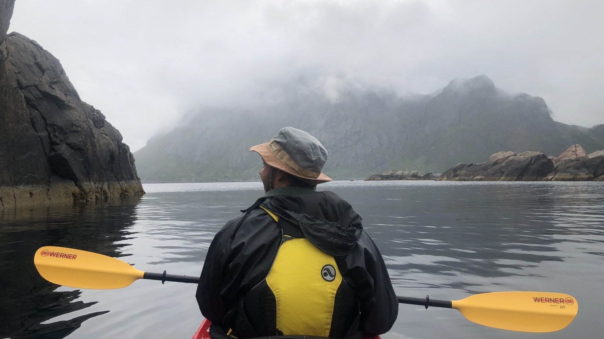 Person kayaking on misty lake between rocky cliffs with mountains in background.
