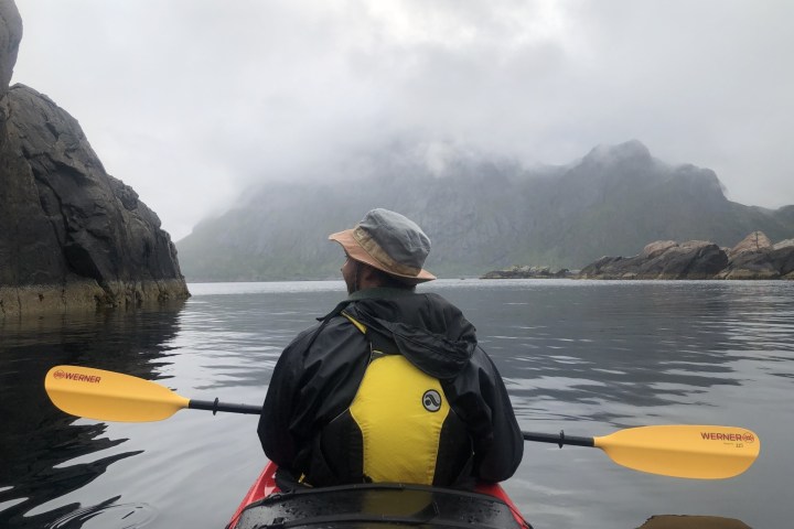 Person kayaking on misty lake between rocky cliffs with mountains in background.