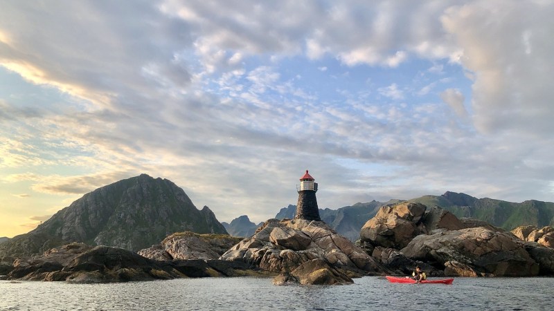 Kayaker in red kayak near rocky shoreline with lighthouse and mountains under cloudy sky.