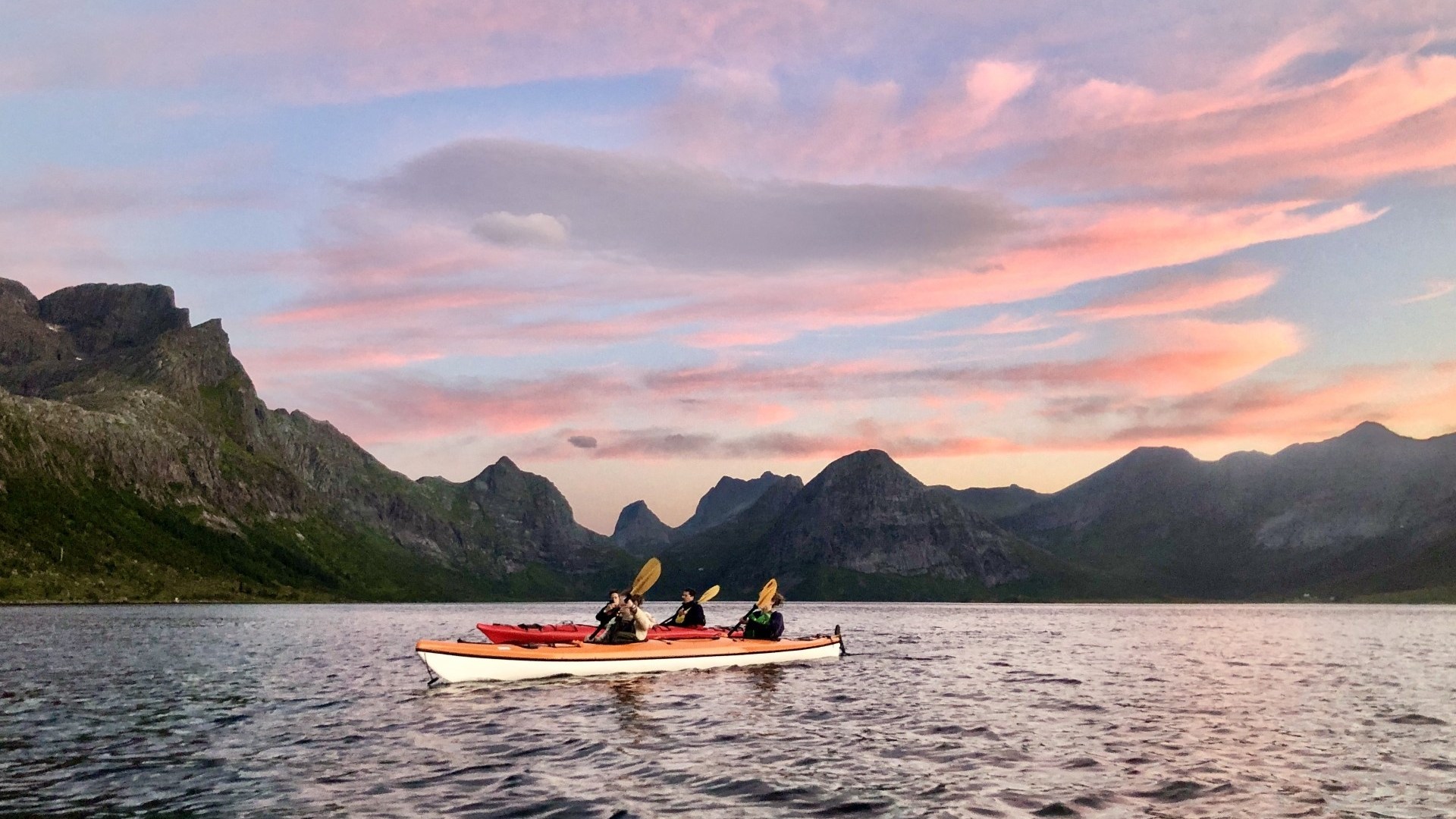 Two kayakers on a lake with mountain backdrop and pink sunset sky.