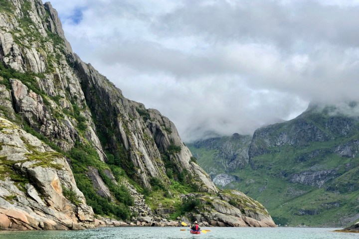 Person kayaking on a calm lake surrounded by steep, green rocky mountains under a cloudy sky.