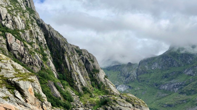 Person kayaking on a calm lake surrounded by steep, green rocky mountains under a cloudy sky.