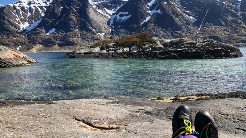 Person's feet on rock facing a clear lake and snow-capped mountains.