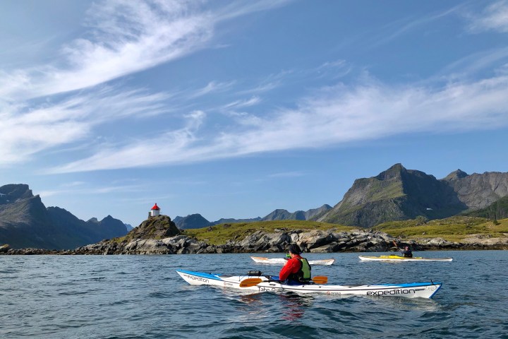 Kayakers on a lake with mountains and a small lighthouse in the background under a blue sky.