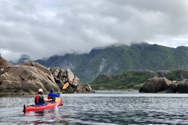 Two people in a kayak paddling near rocky cliffs with green mountains in the background.